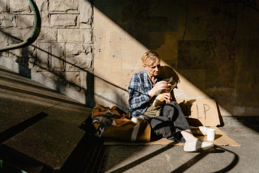 A homeless person sitting under a bridge with a blanket and a baguette.